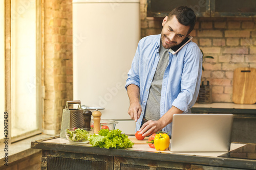 It's so delicious! Casual happy young man preparing salad at home in loft kitchen, using phone, laptop and smiling.