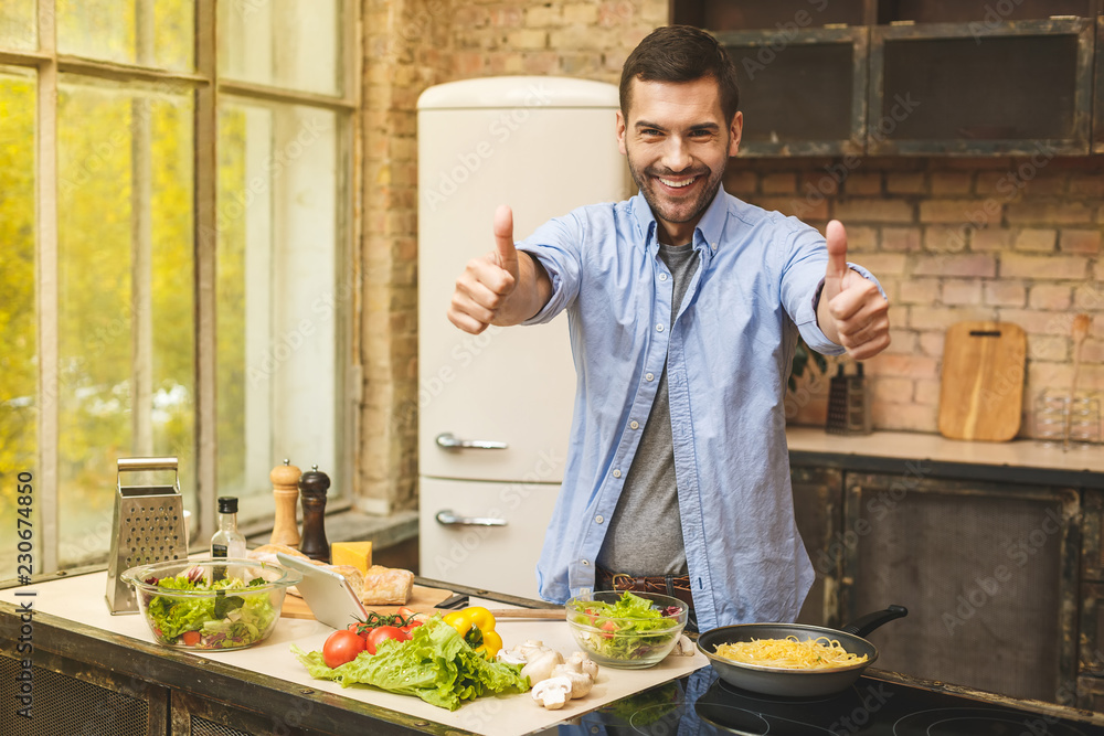 Man preparing delicious and healthy food in the home kitchen on a sunny day. Using tablet computer for searching recipes. Thumbs up. Man preparing delicious and healthy food in the home kitchen on a sunny day. Using tablet computer for searching recipes. Thumbs up.