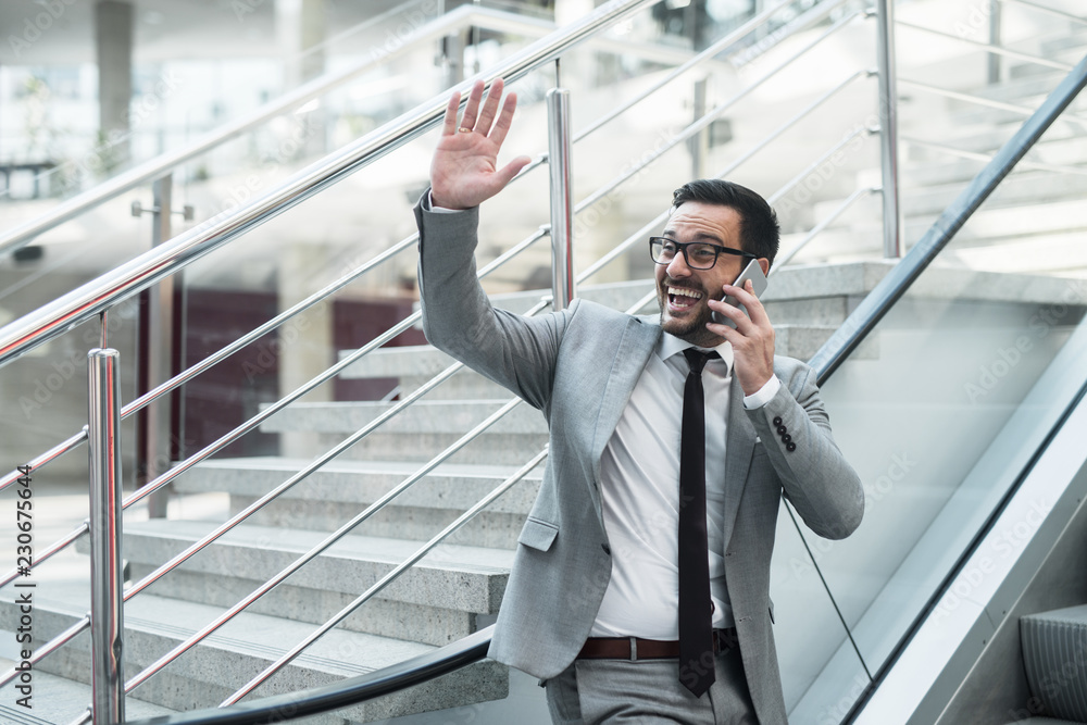 Businessman talking on the smart phone and waving to the colleague while going up the moving stairs. Businessman talking on the smart phone and waving to the colleague while going up the moving stairs.