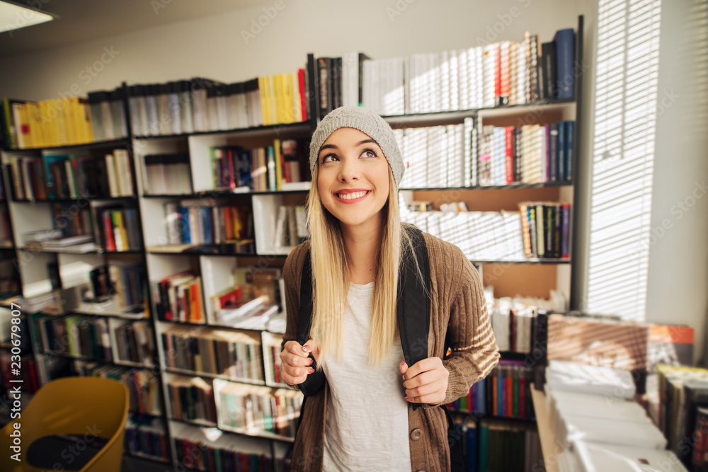Cute happy school girl standing in library with backpack on her back. Smiling and looking away and satisfied. Cute happy school girl standing in library with backpack on her back. Smiling and looking away and satisfied.