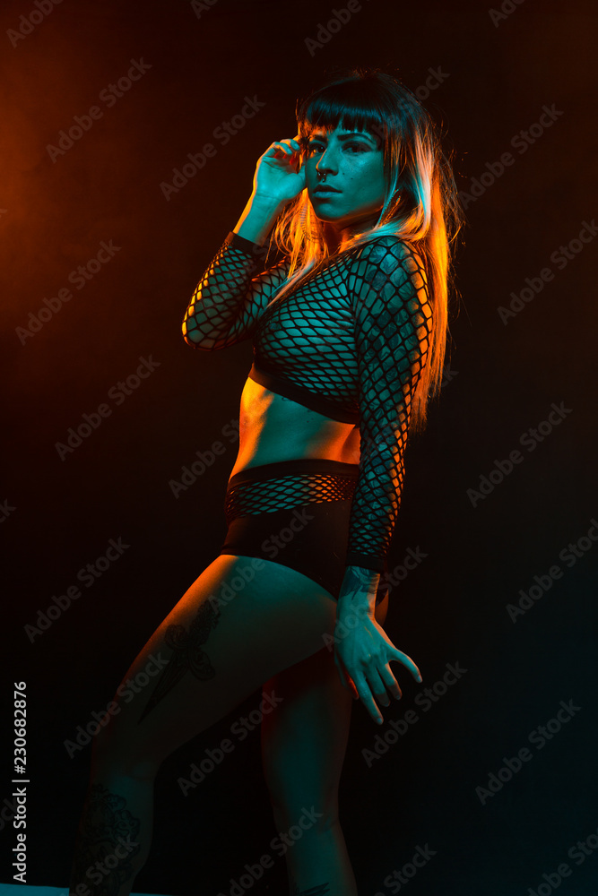 Alternative model with bangs and colored hair poses under teal and orange light wearing a fishnet top Alternative model with bangs and colored hair poses under teal and orange light wearing a fishnet top