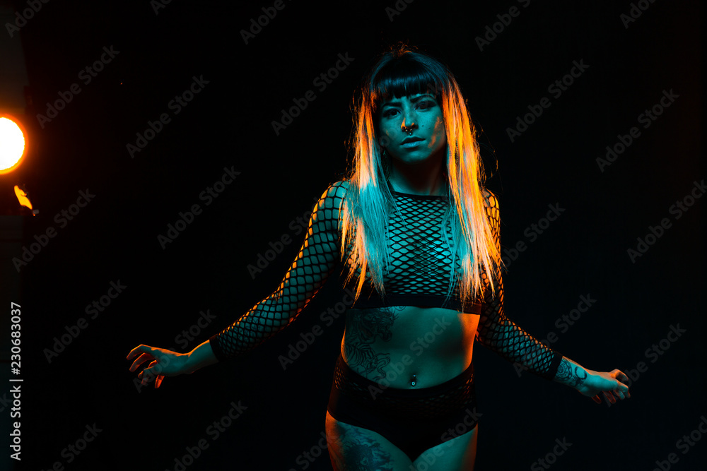 Alternative model with bangs and colored hair poses under teal and orange light wearing a fishnet top Alternative model with bangs and colored hair poses under teal and orange light wearing a fishnet top
