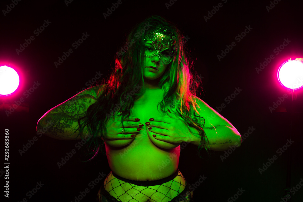 Curvy alternative model with colored hair and fur coat and fishnets under green and pink lighting