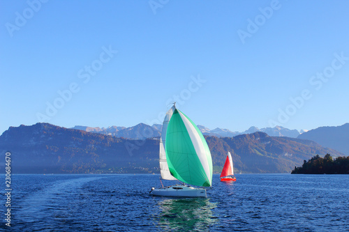 Active rest on clear sunny day. Yachts with red and green sail on lake surrounded by mountains.