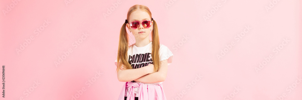 Girl with red tails on a pink background. A charming girl in bright sports clothes and pink glasses folded her arms on her chest.