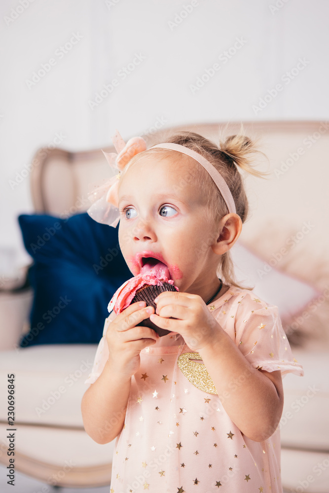 children's birthday. funny two-year-old Caucasian girl in pink dress standing to bedroom of house the background of couch and eating, holding cake dessert. Face and mouth smeared, smeared with cream children's birthday. funny two-year-old Caucasian girl in pink dress standing to bedroom of house the background of couch and eating, holding cake dessert. Face and mouth smeared, smeared with cream