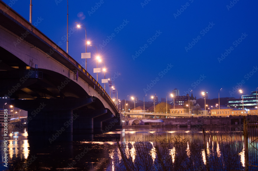 Night city landscape with bridge over river and beautiful glowing lanterns on quay.