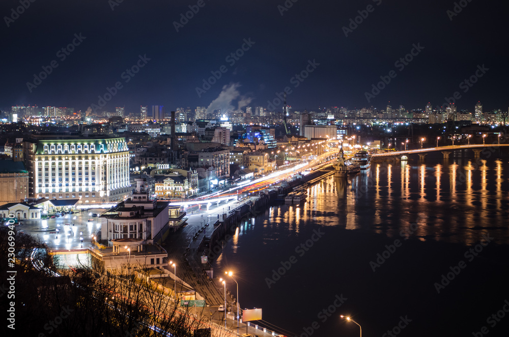 Panorama of night city landscape with river and bridge. Reflection of glowing lanterns in water.