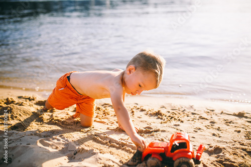 Caucasian child boy playing toy red tractor, excavator on a sandy beach by the river in red shorts at sunset day