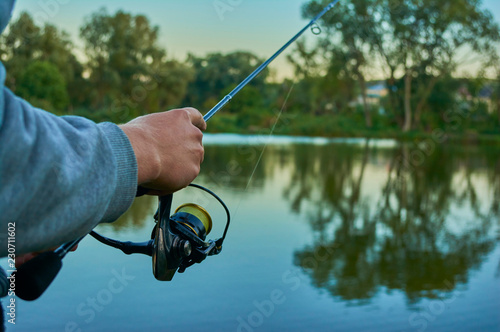 Lake at sunset. A fisherman on the bank. Fishing rod wheel closeup. Spinning reel. The concept of outdoor activities. Tackles for pike, perch, zander.