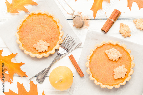 Pumpkin pie with cinnamon and cookies on gray napkins on white wooden background with autumn yellow leaves.