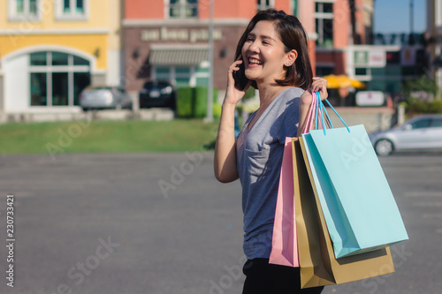 Beautiful young shopaholic Asian woman using smartphone for talking while she walking to buy cosmetics, clothes etc. with shopping bags in their hands. Lifestyle woman enjoy her holidays concept.