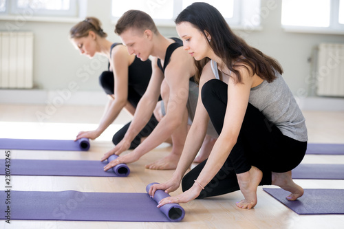 Three young people unrolling purple mats, millennial girls and guy wearing sportswear barefoot unwrap personal individual rubber carpets preparing for yoga class fitness workout at gym centre studio