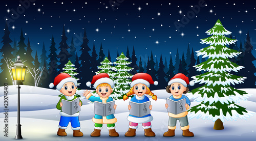 Children singing christmas carols in winter chlotes and santa hat with snowy pine background photo