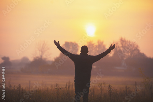 A happy man with hands in the air standing in the field in the early morning and looking at a sunrise