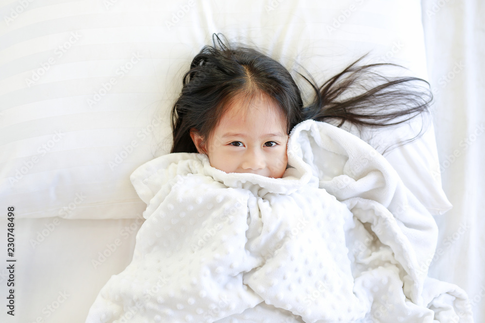 Happy little girl lying on bed with blanket and looking at camera. Top view. Happy little girl lying on bed with blanket and looking at camera. Top view.