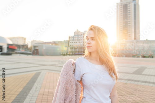 Street portrait of a girl who poses on a camera with a pink coat in her hand and looks sideways on the background of a beautiful sunset.Attractive girl at sunset