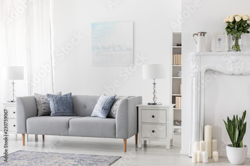 Lamp on white cabinet next to grey couch in simple flat interior with plants and candles. Real photo