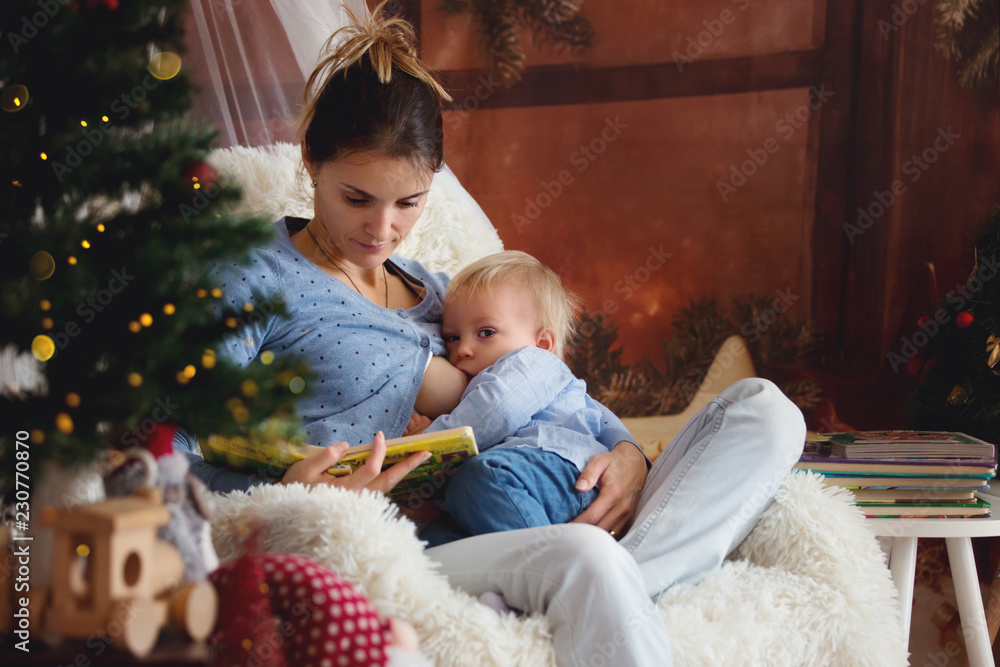 Mother breastfeeding her toddler son sitting in cozy armchair near Christmas tree Mother breastfeeding her toddler son sitting in cozy armchair near Christmas tree