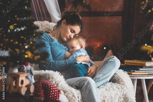 Mother breastfeeding her toddler son sitting in cozy armchair near Christmas tree
