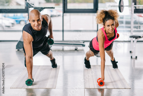 cheerful african american couple of athletes exercising with dumbbells on fitness mats at gym