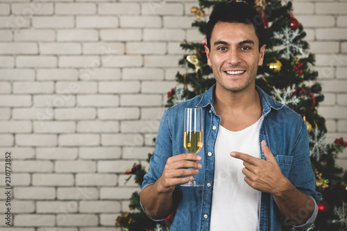 The happy caucasian man is enjoying  christmas with  his gorgeous smile on his face,  pointing to the glass of white wine with the glammorous christmas tree with a copyspace behind
