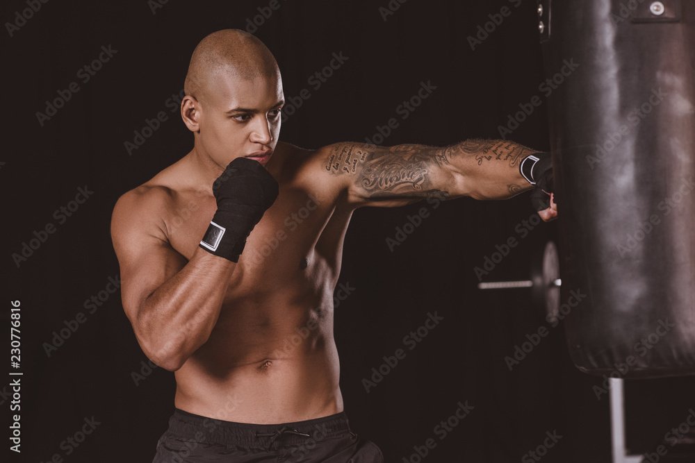 tattooed african american male boxer in boxing bandage training with punching bag at gym