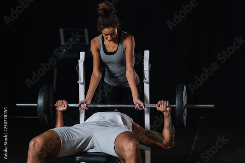 african american woman helping boyfriend to exercising with barbell at gym
