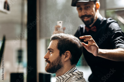 Mustachioed barber dressed in a black shirt and a red bow tie is doing the hair styling to the stylish man sitting in the armchair in a barbershop