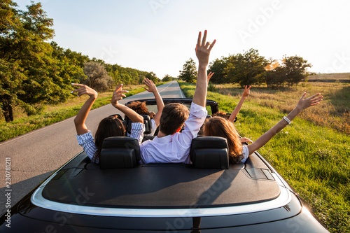 Black cabriolet is on the country road. Company of young girls and guys are sitting in the car hold their hands up on a sunny day.