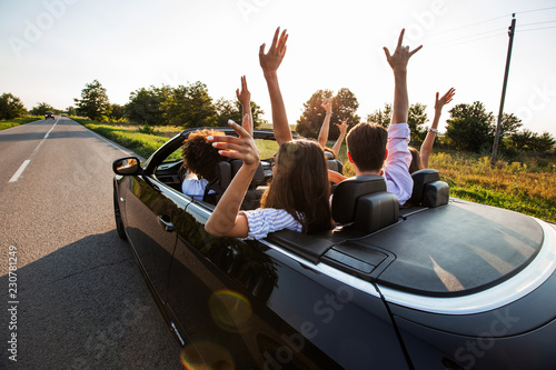 Black cabriolet is on the country road. Happy group of young girls and guys are sitting in the car hold their hands up on a sunny day.