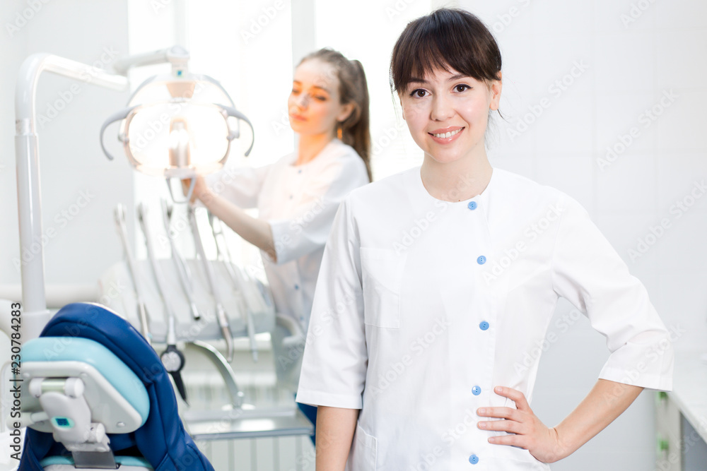 Fototapeta premium Two girls dentists at the workplace, the dentist in the background preparing equipment for the reception of the patient