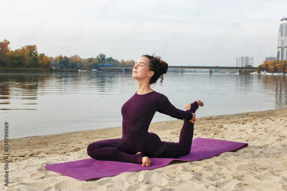 A young girl practices yoga by the river. Exercises against the background of the river, the shore and the bridge.