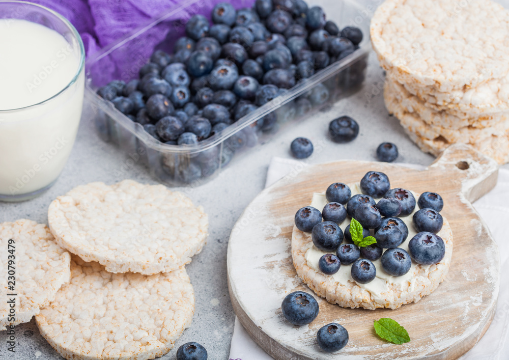 Healthy organic rice cakes with ricotta and fresh blueberries and glass of milk on light stone kitchen background. Top view. Plastic tray of strawberries Healthy organic rice cakes with ricotta and fresh blueberries and glass of milk on light stone kitchen background. Top view. Plastic tray of strawberries