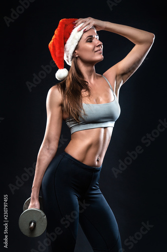 Attractive fitness woman is standing with dumbbell in christmas hat