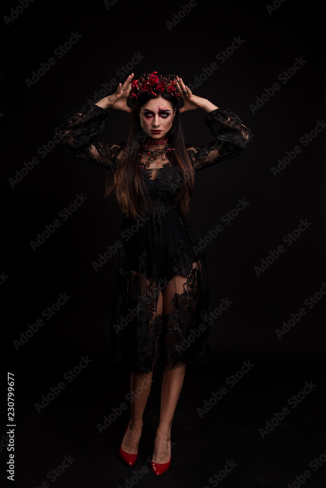 Portrait of a girl in the image for Halloween. Black Widow. Wreath of roses. Makeup for halloween. Eyes like gimlets. Hands at head. Black transparent lace dress. Black background. Portrait of a girl in the image for Halloween. Black Widow. Wreath of roses. Makeup for halloween. Eyes like gimlets. Hands at head. Black transparent lace dress. Black background.