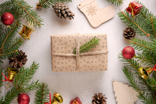 Christmas gift wrapped in craft paper and twine decorated with green spruce twig on white background among pine cones, spruce branches and red and golden decorations. Flat lay style
