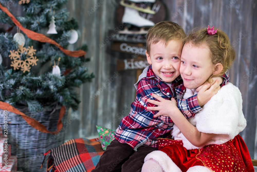 Φωτογραφία Two small children, brother and sister, are sitting on the background of the Christmas interior and gifts