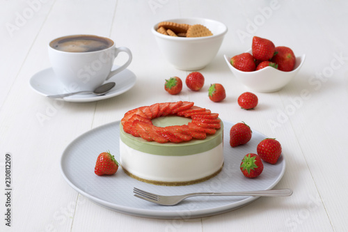 Cooking demo. No-bake two-layered mini strawberry matcha cheesecake. Strawberry slices decorate the top of the cake. 3 cups with coffee, cookies and strawberry in background.