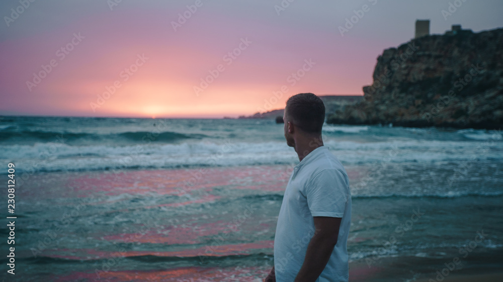 Rear shot of young man in white shirt standing on the beach and looking at the beautiful pink sunset and scenic mediterranean sea with white waves. Dusk. Man silhouette Rear shot of young man in white shirt standing on the beach and looking at the beautiful pink sunset and scenic mediterranean sea with white waves. Dusk. Man silhouette