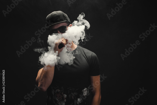 Vape man. Portrait of a handsome young white guy in a modern black cap vaping and letting off puffs of steam from an electronic cigarette 