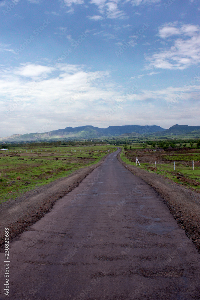 Open dirt road with mountain view, green field and nice cloudy blue sky. Maharashtra, India.