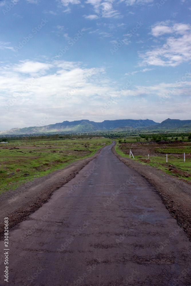 Open dirt road with mountain view, green field and nice cloudy blue sky. Maharashtra, India. Open dirt road with mountain view, green field and nice cloudy blue sky. Maharashtra, India.