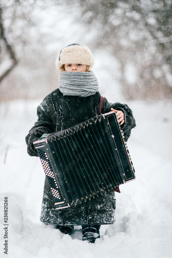Young happy musician. Comic girl holding accordion. Lovely child in funny  winter clothes outdoor lifestyle portrait. Odd unusual female kid wearing  oversized adult padded jacket. Country baby fashion Stock Photo