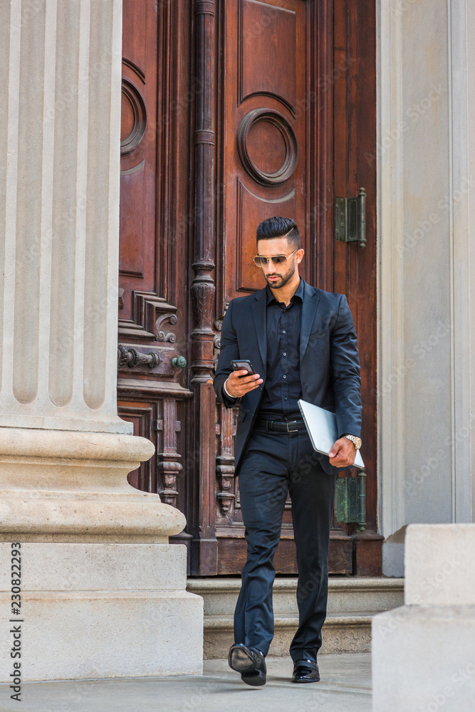Young East Indian American Businessman traveling, working in New York, with beard, wearing black suit, sunglasses, holding laptop computer, walking out from office building, reading on cell phone