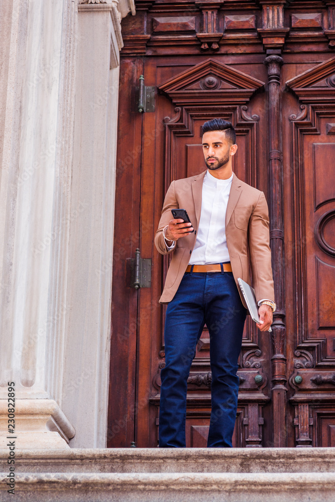 Young East Indian American Man with beard, wearing light brown blazer,  white round collar shirt, blue pants, holding laptop computer, standing in  front old style door way, texting on cell phone.. Stock