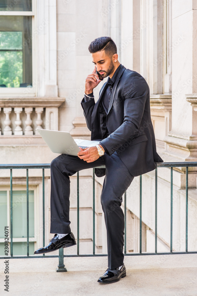 Young East Indian American Businessman with beard working in New York, wearing black suit, leather shoes, sitting on railing inside office building, working laptop computer, talking on cell phone..