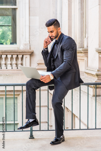 Obraz na płótnie Young East Indian American Businessman with beard working in New York, wearing black suit, leather shoes, sitting on railing inside office building, working laptop computer, talking on cell phone.. w salonie