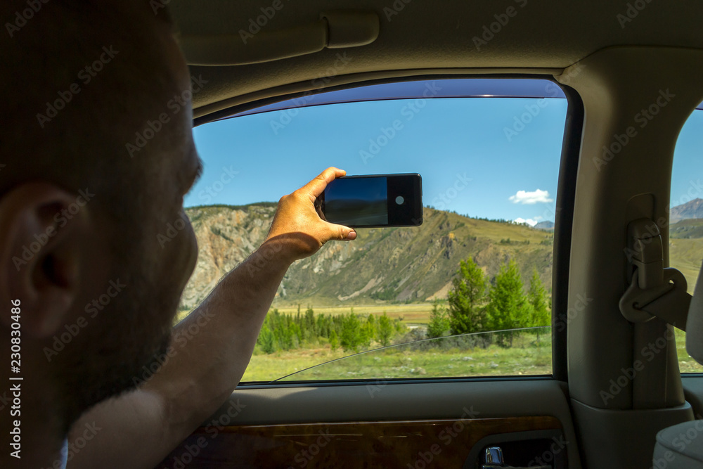 A young man pulling one hairy hand into the open window of a moving car holds a smartphone taking a photo of the mountains in the Altai with blue clear sky on an autumnal summer day during a journey