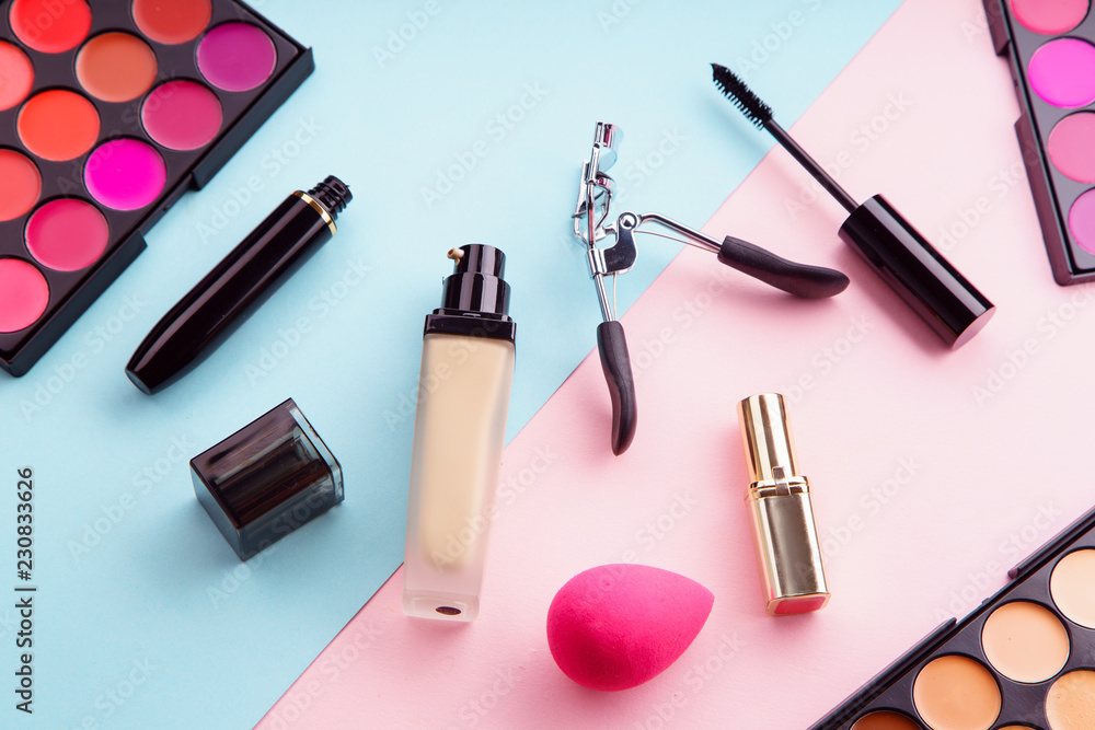 Top view picture of makeup products: lipstick and concealer palettes, foundation, mascara and eyelash curler on pink and blue background. Make-up artist kit concept Top view picture of makeup products: lipstick and concealer palettes, foundation, mascara and eyelash curler on pink and blue background. Make-up artist kit concept